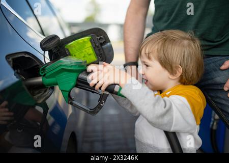 Süßer kleiner blonder Junge, der eine Pumpdüse hält. Kleiner, lustiger Junge, der Vater hilft, das Auto an einer Tankstelle zu tanken. Papas kleiner Helfer. Stockfoto