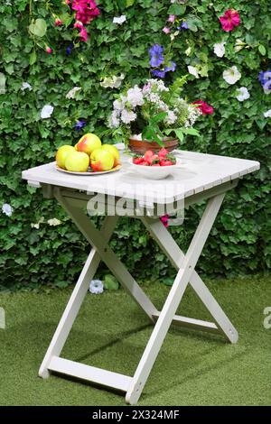 Äpfel, Erdbeeren und Blumen auf weißem Tisch neben grüner Hecke. Stockfoto