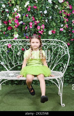 Das kleine süße Mädchen sitzt auf einer Bank und schwingt die Beine in der Nähe einer grünen Hecke. Stockfoto