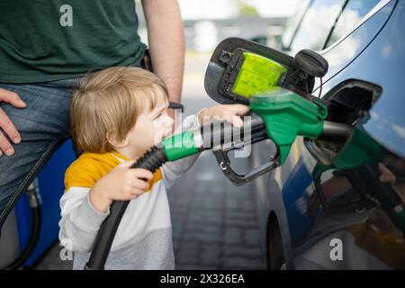 Süßer kleiner blonder Junge, der eine Pumpdüse hält. Kleiner, lustiger Junge, der Vater hilft, das Auto an einer Tankstelle zu tanken. Papas kleiner Helfer. Stockfoto