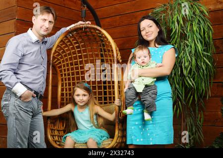 Das kleine Mädchen sitzt im Hängesessel und Vater, Mutter mit Baby steht neben einem Baum im Topf. Stockfoto