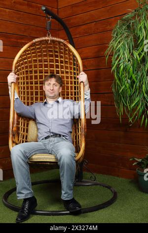 Glücklicher Mann sitzt in einem Korbstuhl neben der hölzernen Hauswand. Stockfoto