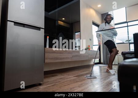 Junge Afroamerikaner, die zu Hause Spaß mit Mopp und Musik haben, Tanzbewegungen zeigen und im Wohnzimmer singen. Afroamerikaner, der Holz wischt Stockfoto
