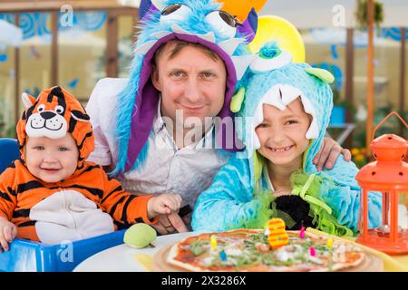Vater mit Tochter in Monsterkostümen und kleiner Junge im Tigerkostüm feiern den Geburtstag in einem Café Stockfoto