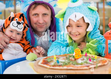 Vater mit Tochter in Monsterkostümen und kleiner Junge im Tigerkostüm feiern den Geburtstag in einem Café mit Pizza Stockfoto