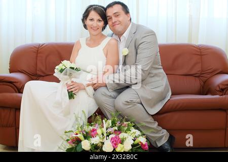 Schöne Braut und Bräutigam sitzen auf der Couch mit einem Korb voller Blumen zu den Füßen Stockfoto