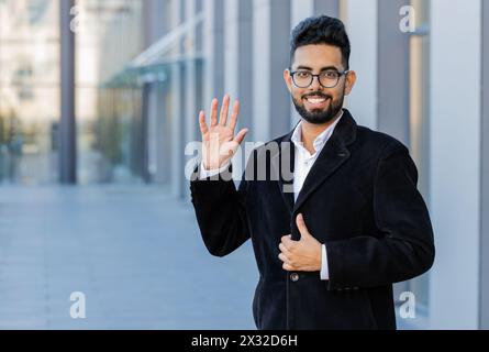 Hallo. Indischer Geschäftsmann lächelt freundlich vor der Kamera, winkende Hände, die hallo, Begrüßung oder Abschied heißen mit gastfreundlichem Ausdruck im Freien willkommen. Ein arabischer Hindu-Freelancer in der Innenstadt Stockfoto
