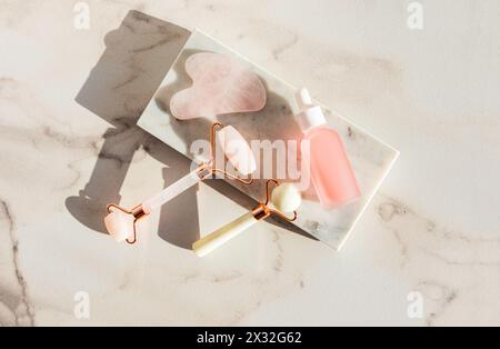 Zwei verschiedene Massageroller, Gua Sha, Pipette mit Naturpflegemittel, Serum oder Öl auf Marmorpodium und Hintergrund. Draufsicht. Sonnig. Stockfoto