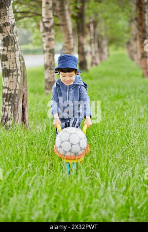 Der kleine Junge trägt eine Plastikkarre mit Ball im Park Stockfoto