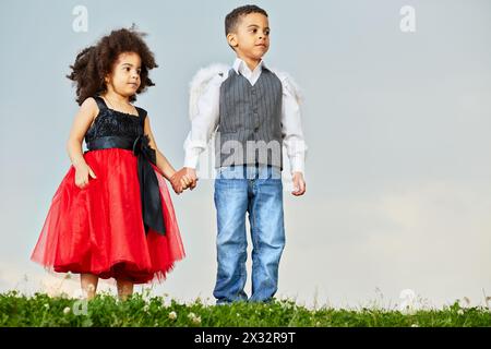 Das kleine Mädchen und der Junge stehen auf einer grasbewachsenen Wiese vor bewölktem Himmel, das Mädchen in schwarz-rotem Kleid, der Junge in grauem Anzug mit Weste und Engelsflügeln auf dem Rücken Stockfoto