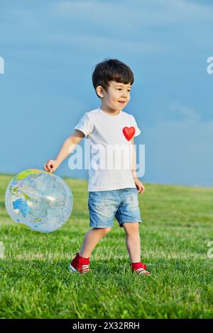 Der kleine Junge in Shorts und weißem T-Shirt mit rotem Herz geht auf eine grasbewachsene Wiese und hält aufblasbare Kugelkugel in der Hand Stockfoto