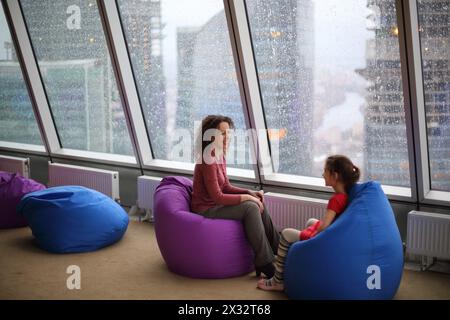 Frau, die mit ihrer Tochter spricht und an einem regnerischen Tag auf gepolsterten Hockern an der Glaswand sitzt Stockfoto