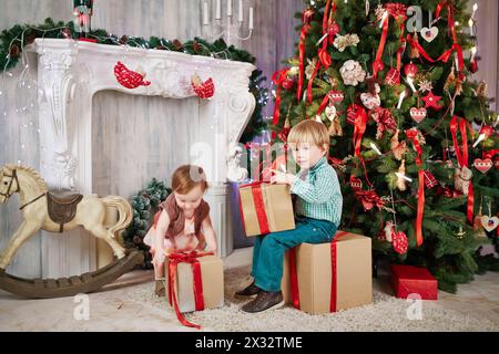 Der kleine Junge sitzt auf einer großen Geschenkbox aus Karton, hält eine andere in der Hand neben einem geschmückten Weihnachtsbaum, das kleine Mädchen versucht, die Schachtel zu nehmen Stockfoto