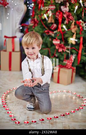 Der kleine Junge steht auf dem Boden auf einem Knie zwischen brennenden Teelichtern und hält eines auf seiner Palme unter dem Weihnachtsbaum Stockfoto