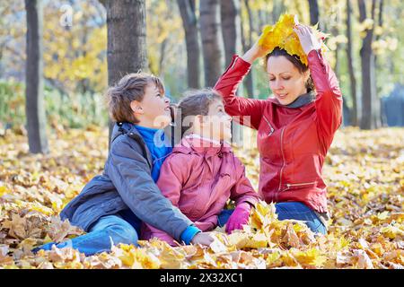 Kinder sehen ihre Mutter an, die nebenan sitzt und eine Krone aus gelben Ahornblättern anprobiert Stockfoto