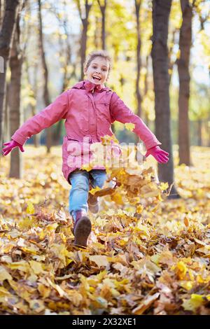 Das kleine Mädchen rennt durch den Herbstpark und wirft mit ihren Stiefeln gefallene Blätter auf Stockfoto