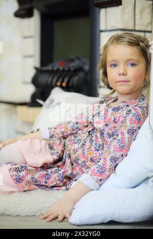 Das kleine süße Mädchen liegt auf Pelz und Kissen in der Nähe des Kamins und schaut in die Kamera in einem gemütlichen Zimmer. Stockfoto