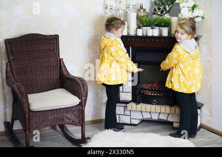 Zwei kleine Mädchen in der gleichen Kleidung stehen neben Kamin und Korbschaukelstuhl in einem gemütlichen Zimmer. Konzentriere dich auf das richtige Mädchen. Stockfoto