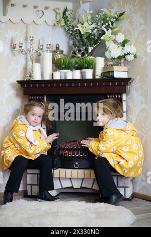 Zwei kleine Mädchen in gleicher Kleidung sitzen neben Kamin und Korbschaukelstuhl und warme Hände in einem gemütlichen Zimmer. Konzentriere dich auf das richtige Mädchen. Stockfoto