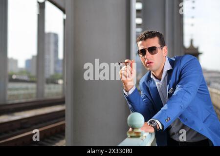 Schöner Mann in blauer Jacke und Sonnenbrille mit Zigarren steht auf der Brücke. Stockfoto