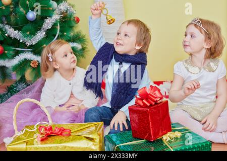 Drei Kinder sitzen auf einem Pelzteppich unter dem weihnachtsbaum, der Junge klingelt die Handglocke, die er in seiner Hand nach oben gestreckt hält Stockfoto