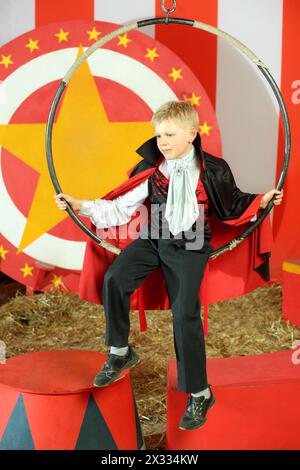 Kleiner Junge in schwarzem Anzug und rotem Mantel, der auf einem Zirkusring sitzt Stockfoto