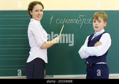 Junge und Lehrer mit Zeiger stehen in der Nähe der Tafel mit Inschrift 1. September im Klassenzimmer der Schule. Fokus auf Frau. Stockfoto