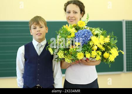 Glücklicher Lehrer mit großen Blumen und Jungen stehen neben der Tafel in der Schule. Konzentriere dich auf den Jungen. Stockfoto