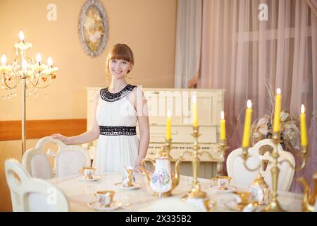 Happy Girl steht neben einem klassischen weißen Tisch mit Geschirr und beleuchteten Kerzen. Stockfoto
