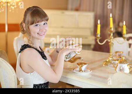 Hübsches Mädchen sitzt an einem klassischen weißen Tisch mit Geschirr und brennenden Kerzen und gießt Tee. Stockfoto