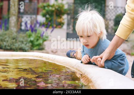 Außenporträt eines entzückenden kleinen Jungen, der draußen spielt, Mutter, die das Kind davon abhält, in den alten, dreckigen Brunnen zu kommen Stockfoto