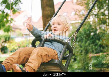 Außenporträt des entzückenden kleinen Jungen, der Spaß auf der Schaukel auf dem Spielplatz hat Stockfoto