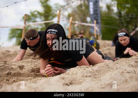 Fröhliche ukrainische Frau krabbelt im Sand unter Stacheldraht während der Spartan Race Challenge in der Ukraine. Kiew - 20. April 2024 Stockfoto