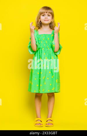 Das Schulmädchen drückte die Daumen und schaute in die Kamera, fragte nach Glücksnachrichten, wünschte sich gute Prüfungsergebnisse und träumte vom Sieg im Spiel. Kind vor einem Kind, isoliert auf gelbem Hintergrund. Vertikal Stockfoto