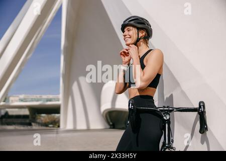 Lächelnde Radfahrerin legt Helm auf den Kopf, bevor sie in der Stadt mit dem Fahrrad fahren geht Stockfoto