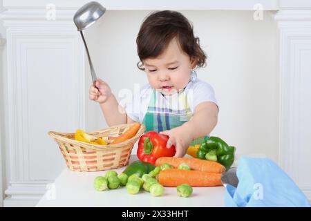 Kleiner Junge in der Küchenschürze mit Kelle und Gemüse am Tisch Stockfoto