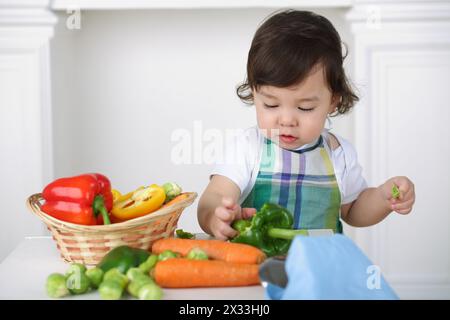 Kleiner Junge in der Küchenschürze, der mit Gemüse am Tisch spielt Stockfoto