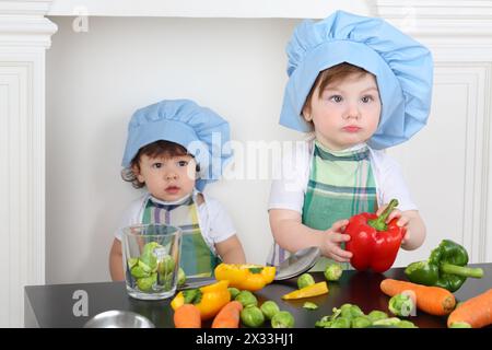 Kleines Mädchen in der Küchenschürze und Mütze mit großem roten Pfeffer und einem kleinen Jungen Stockfoto