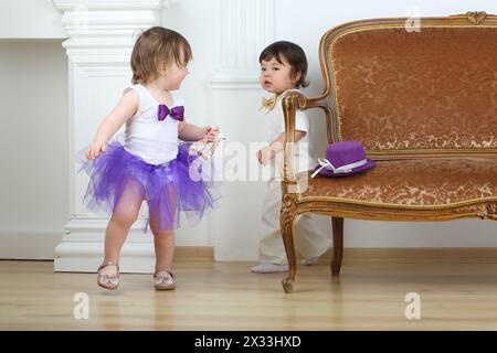 Kleines Mädchen im lila Rock, das vor dem Jungen im weißen Anzug um das Sofa läuft Stockfoto