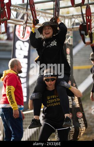 Fröhliches junges Mädchen, das beim Spartan Race Competition auf dem Rücken ihres Freundes sitzt. Kiew - 20. April 2024 Stockfoto