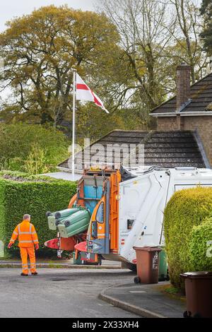 Recyclingarbeiter in orangefarbenen Jacken und Hosen laden Müll aus grünen Mülltonnen in einen Zerkleinerungswagen. Stockfoto