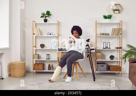 Frau mit gebrochenem Bein, die auf einem Stuhl sitzt und ihr Handy mit einem überraschten Gesicht ansieht Stockfoto