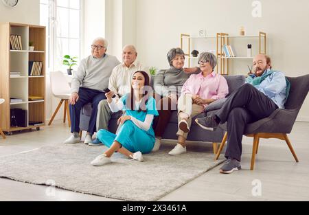 Gruppe verschiedener Senioren, die im Altersheim fernschauen, mit einer jungen Krankenschwester in der Ferne. Stockfoto