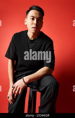 Ein stilvoller asiatischer Mann in schwarzer Kleidung sitzt auf einem Hocker vor einer auffälligen roten Wand in einem Studio-Setting. Stockfoto