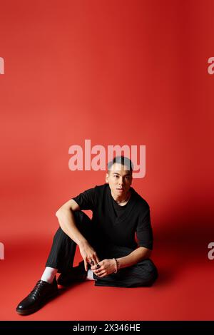 Ein stilvoller asiatischer Mann in schwarzer Kleidung sitzt anmutig auf dem Boden vor einem auffälligen roten Hintergrund. Stockfoto