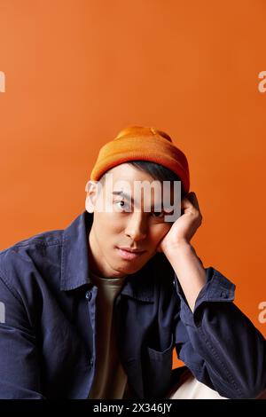 Hübscher asiatischer Mann in einem blauen Hemd und einem orangefarbenen Hut, der selbstbewusst vor einem leuchtend orangen Hintergrund in einem Studio steht. Stockfoto