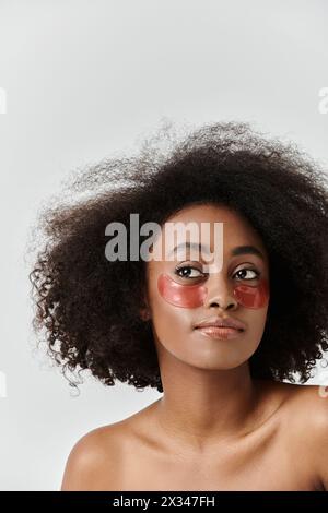 Eine auffällige afroamerikanische Frau mit lockigem Haar, die ein leuchtendes rotes Augenfleck trägt und ein einzigartiges und auffälliges Hautpflegekonzept präsentiert. Stockfoto