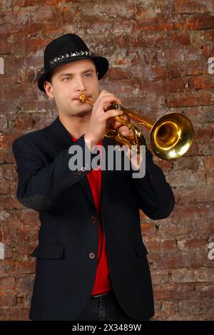 Junger, gutaussehender Mann im Anzug und Hut spielt Trompete im Studio mit Ziegelwand Stockfoto