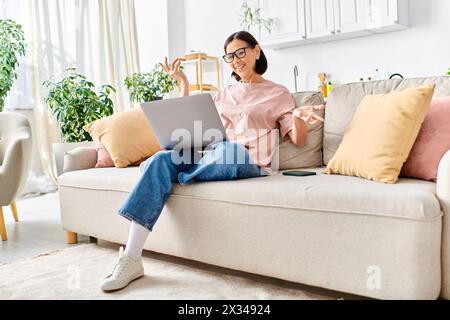 Eine reife Frau in gemütlichen Haushaltswaren sitzt mit einem Laptop auf einer Couch. Stockfoto