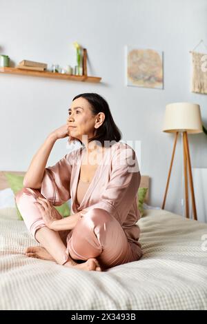 Eine reife Frau in kuscheliger Hauswäsche sitzt friedlich auf einem Bett neben einer glühenden Lampe. Stockfoto